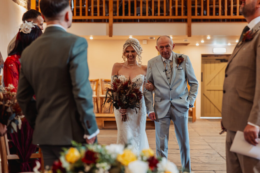 A smiling bride in a white dress walks down the aisle at Ashes Barn in Staffordshire, arm-in-arm with an older man in a light suit, approaching guests and the groom in a warmly lit rustic venue beautifully decorated with flowers.
