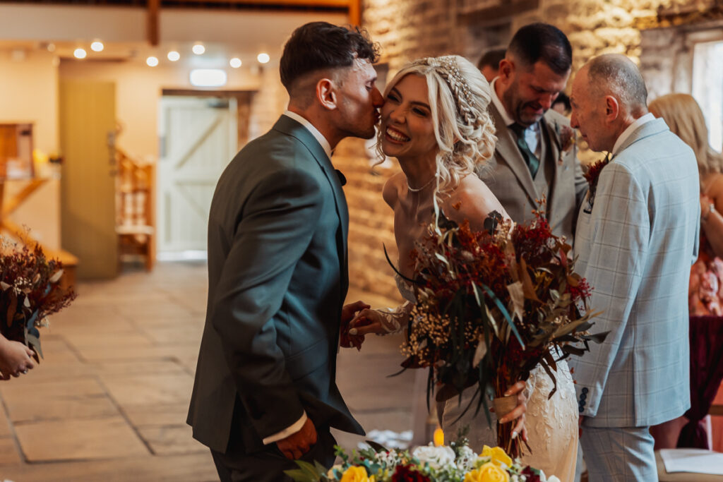 A groom in a green suit leans in to kiss his smiling bride, who holds a bouquet of autumn flowers. Guests in formal attire stand nearby at their Ashes Barn wedding in Staffordshire, as warm indoor lighting highlights the joyful moment.
