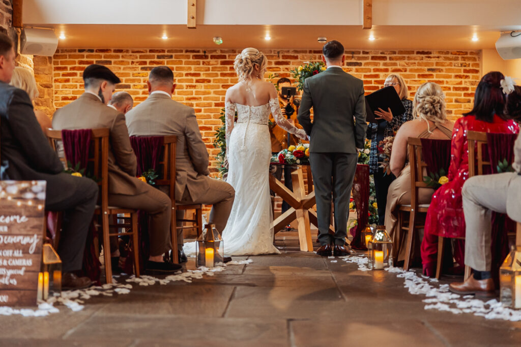 A bride and groom stand at the altar during an indoor wedding ceremony at Ashes Barn in Staffordshire, facing an officiant. Guests are seated on either side, and flower petals line the aisle. Warm lighting and brick walls create a cozy atmosphere.