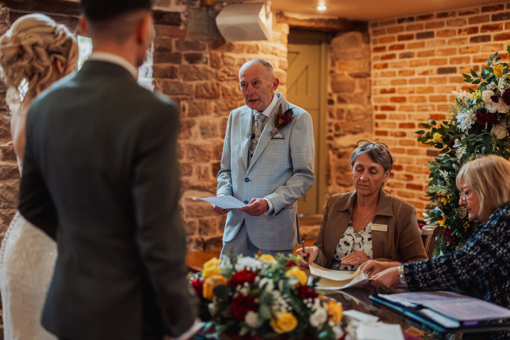 An older man in a light suit stands and speaks, holding papers at a wedding ceremony at Ashes Barn in Staffordshire, while two women sit at a table with documents and flowers. The couple, partially visible, faces the speaker in a warmly lit, rustic room.