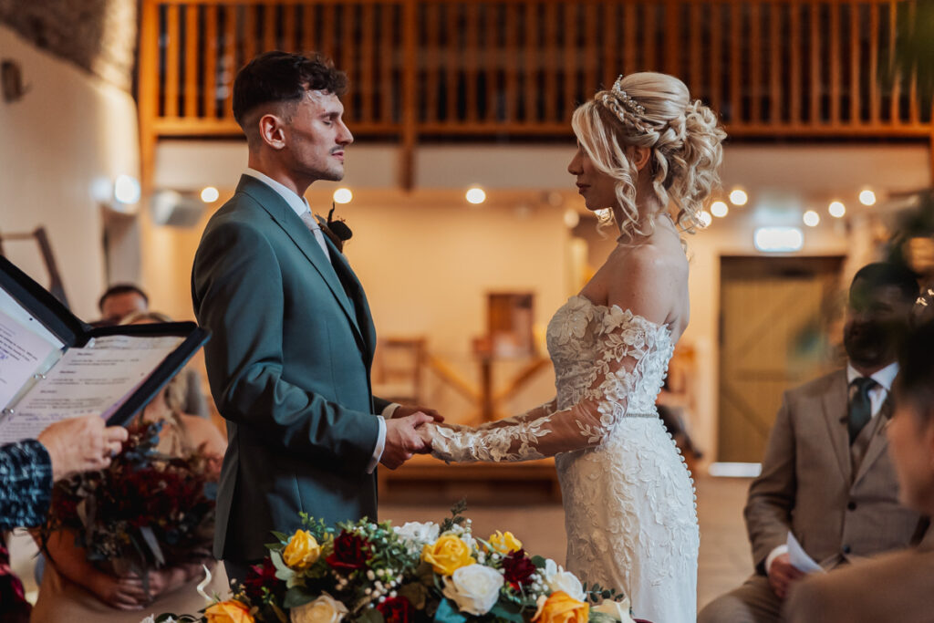 A bride and groom stand facing each other and holding hands during their Wedding ceremony indoors at Ashes Barn, Staffordshire, surrounded by a few seated guests and colorful flower arrangements.
