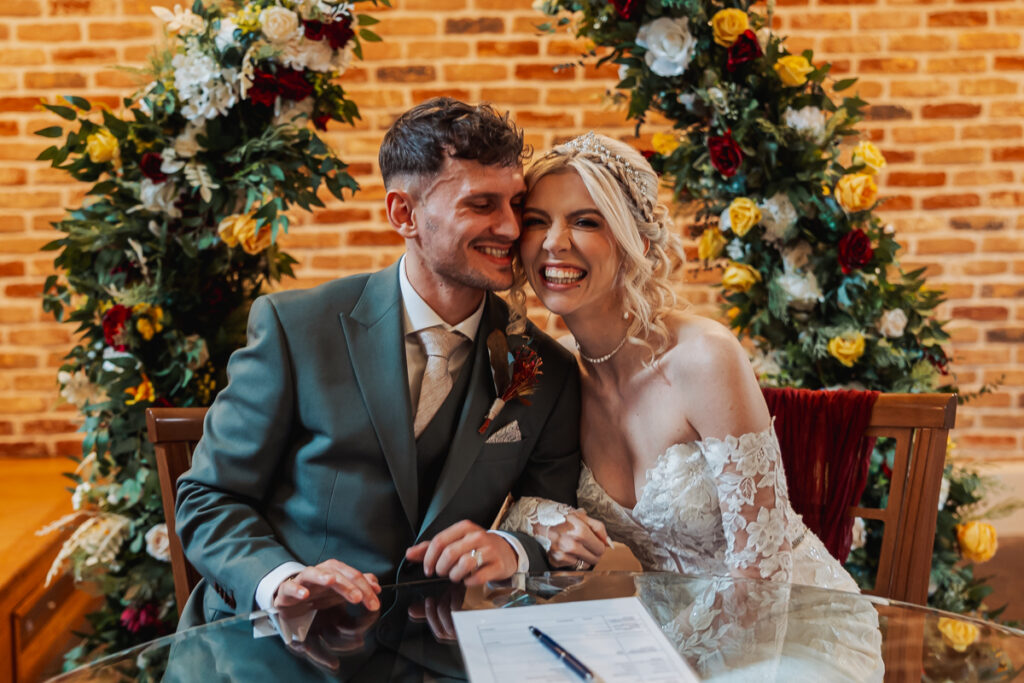 A smiling bride and groom sit close together, holding hands, in front of a decorated brick wall with floral arrangements at Ashes Barn, Staffordshire. A document and pen on the glass table suggest they’ve just signed their wedding papers.