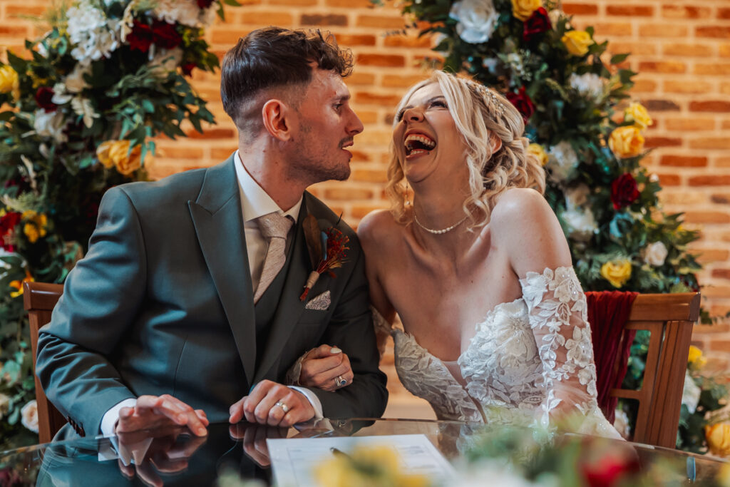 A bride and groom sit arm-in-arm, laughing joyfully together at a table decorated with flowers and greenery, with a brick wall and vibrant floral arrangements in the background at Ashes Barn in Staffordshire.
