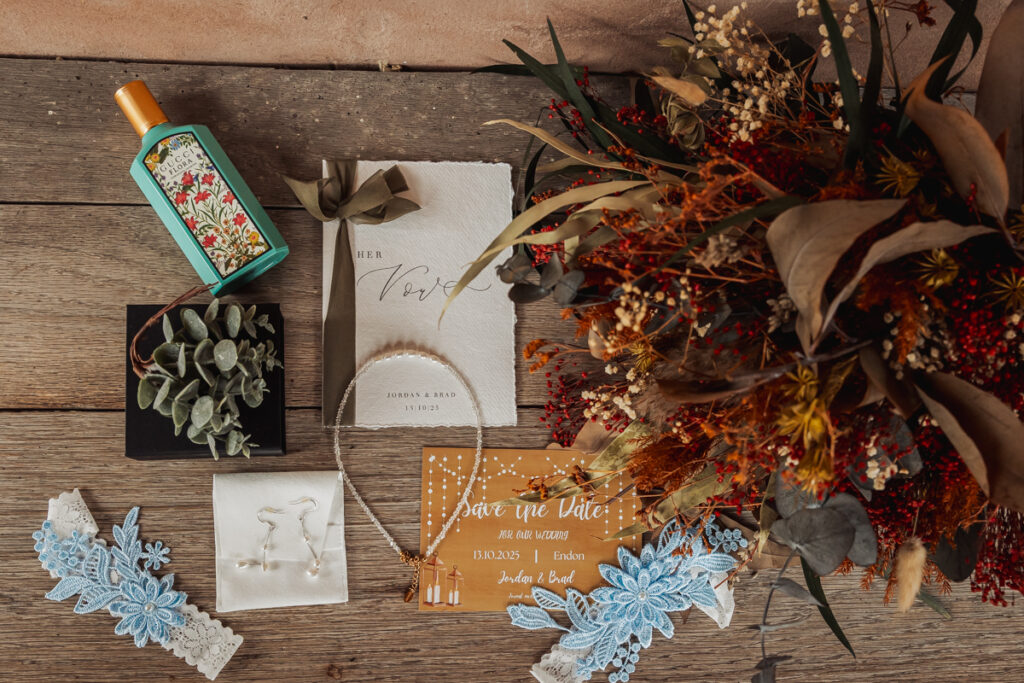 Flat lay of wedding items on a wooden surface, including a bouquet of dried flowers, wedding invitations, a green floral perfume bottle, blue lace garters, earrings, and a small potted succulent—perfect for an Ashes Barn Wedding in Staffordshire.