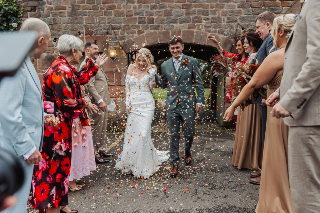A newlywed couple walks outside at Ashes Barn in Staffordshire, smiling, as guests in colorful and neutral outfits throw confetti over them. The festive wedding scene features a stone wall and lanterns in the background.