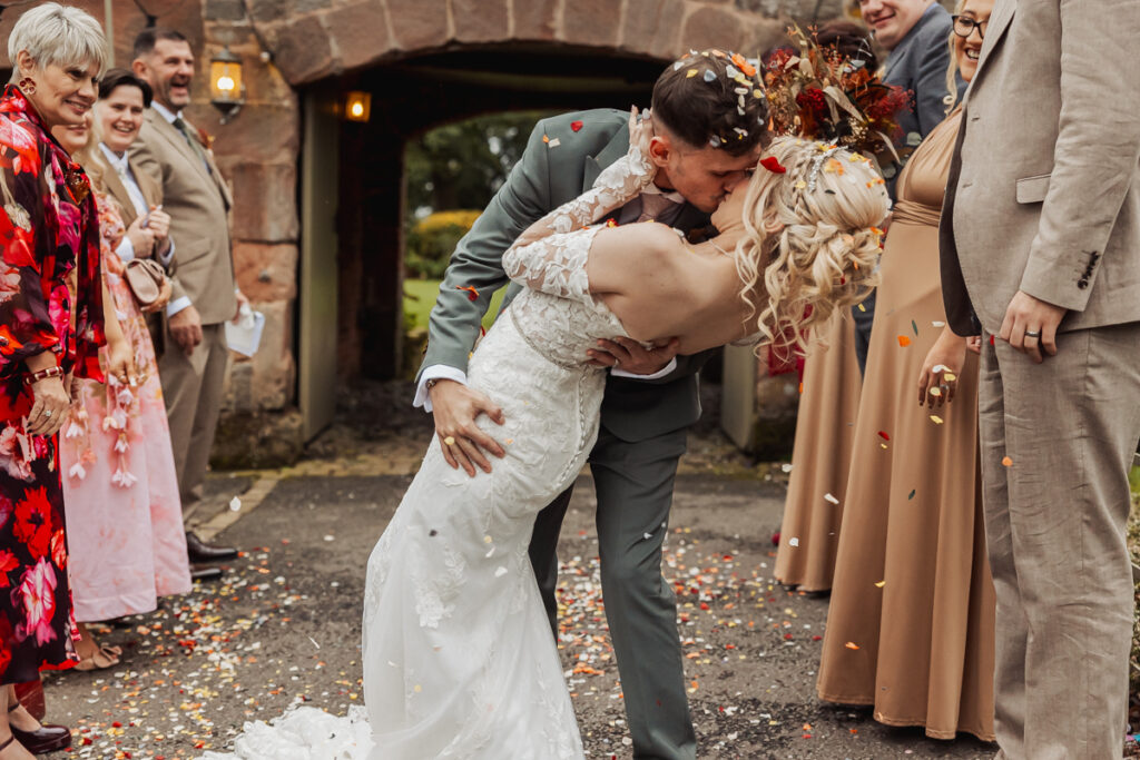 A newlywed couple shares a dramatic kiss as the groom dips the bride at their Ashes Barn Wedding. Guests in formal attire smile and toss flower petals outside the rustic stone building in Staffordshire, creating a festive and cheerful scene.