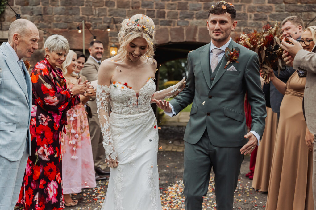 A bride and groom walk hand in hand outdoors at Ashes Barn in Staffordshire, smiling, as guests in colorful outfits throw flower petals and celebrate their wedding. A stone wall is visible in the background.