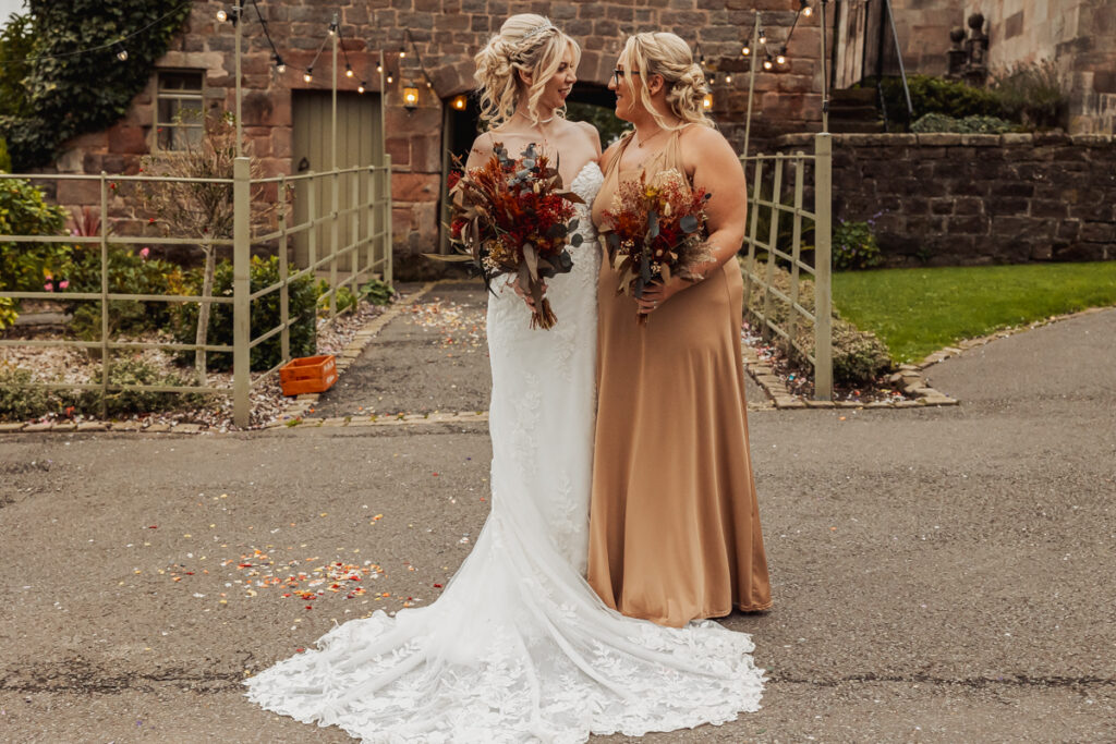 Two women in formal dresses, one in a white bridal gown and the other in a tan dress, smile at each other holding autumn-themed bouquets outside Ashes Barn, a charming wedding venue nestled in the heart of Staffordshire.