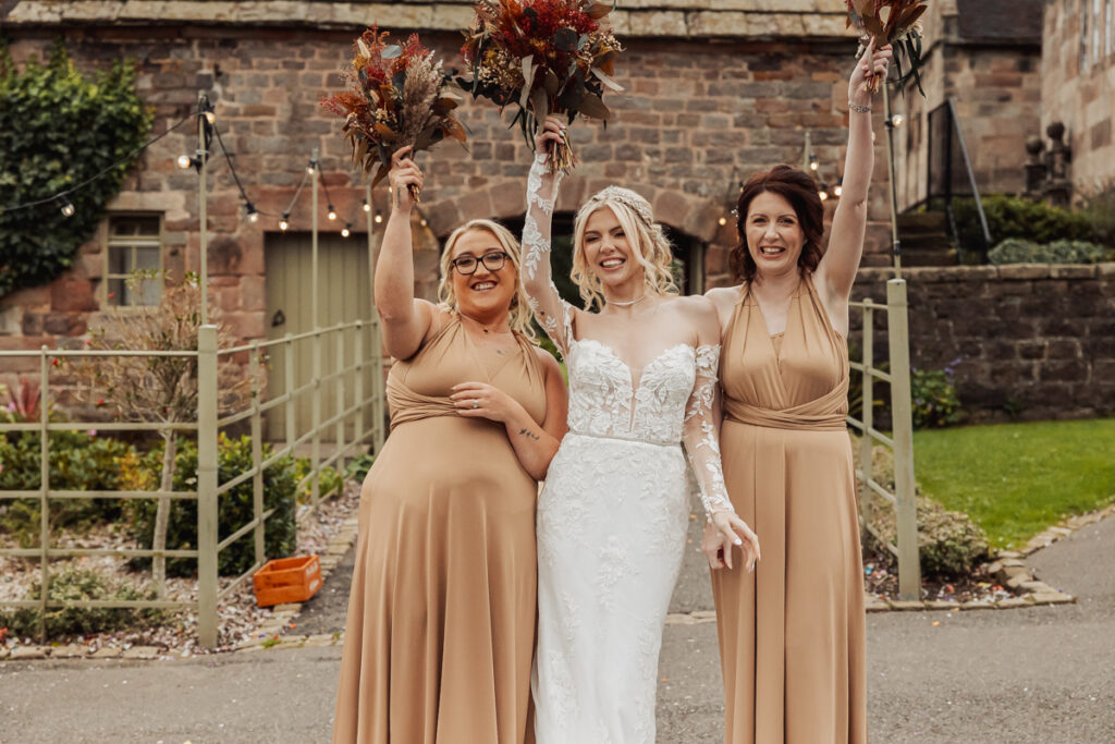 Three women stand smiling outdoors in front of a stone building at Ashes Barn in Staffordshire. The bride, in a white lace gown, is flanked by two bridesmaids in tan dresses. All hold bouquets above their heads, celebrating the wedding.