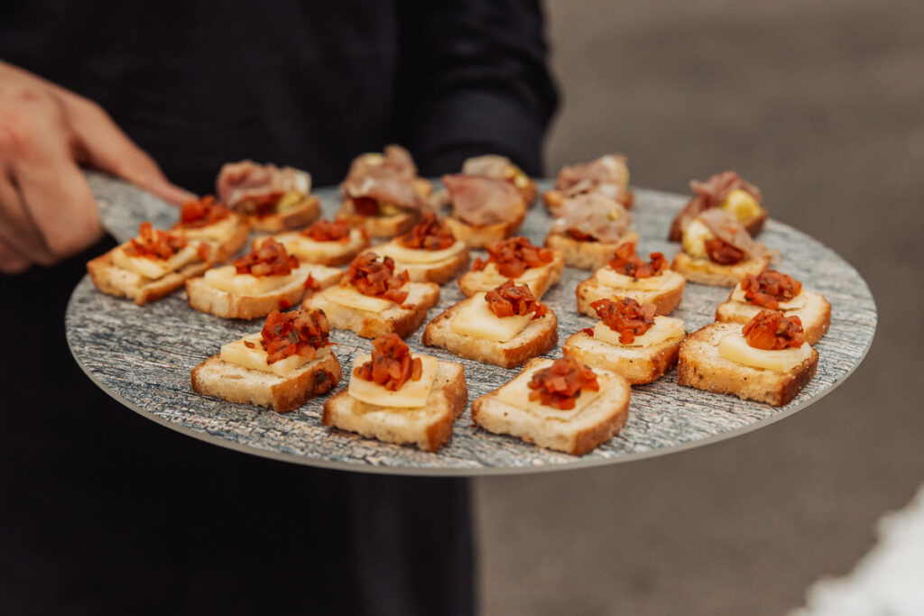 At Ashes Barn in Staffordshire, a person holds a round tray with neatly arranged bite-sized appetizers, including small toasted bread slices topped with cheese and a tomato-based garnish, perfect for a wedding celebration.