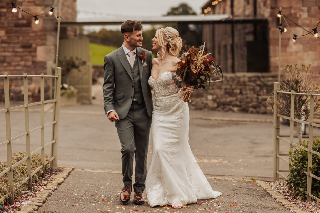 A bride in a white gown holding a bouquet walks arm in arm with her groom in a grey suit. They smile at each other on a path outside Ashes Barn, a rustic Staffordshire wedding venue adorned with string lights.