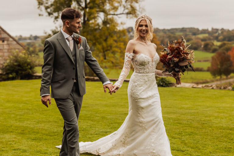 A smiling bride in an off-shoulder lace gown walks hand-in-hand with her groom in a gray suit at Ashes Barn, Staffordshire, their wedding set against grassy lawns, trees, and rolling hills in the background.