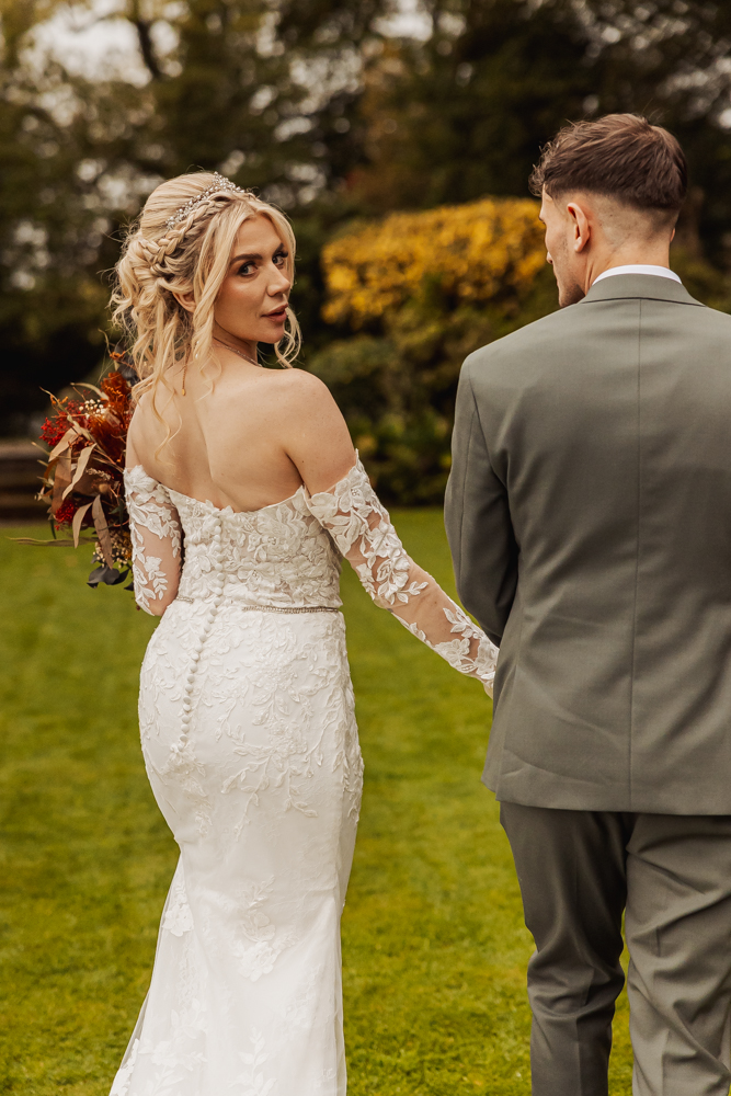 A bride in a white, off-shoulder lace gown holds a bouquet and looks back while walking hand in hand with her groom in a light gray suit on the lush lawn at Ashes Barn, a beautiful wedding venue in Staffordshire.