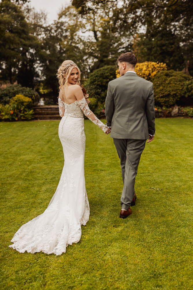A bride in a white lace gown and groom in a gray suit walk hand-in-hand on a green lawn at Ashes Barn, Staffordshire, with the bride smiling back over her shoulder. Lush trees and shrubs surround them in this stunning wedding garden setting.