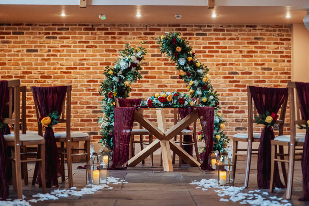 An indoor wedding ceremony setup at Ashes Barn in Staffordshire features wooden chairs draped in burgundy fabric, yellow and orange flowers, lanterns with candles, white petals on the floor, and a floral arch against a brick wall behind a wooden table.