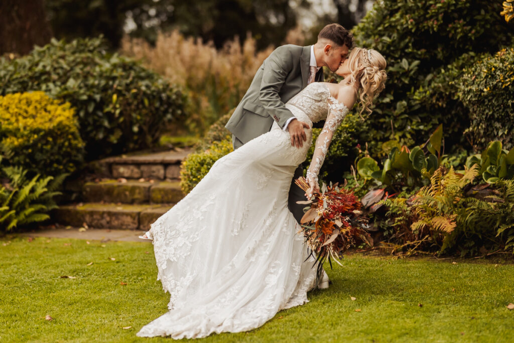 A groom in a suit dips and kisses the bride, who is holding a bouquet and wearing a lace wedding gown, in the lush gardens of Ashes Barn, Staffordshire, surrounded by vibrant greenery on their wedding day.