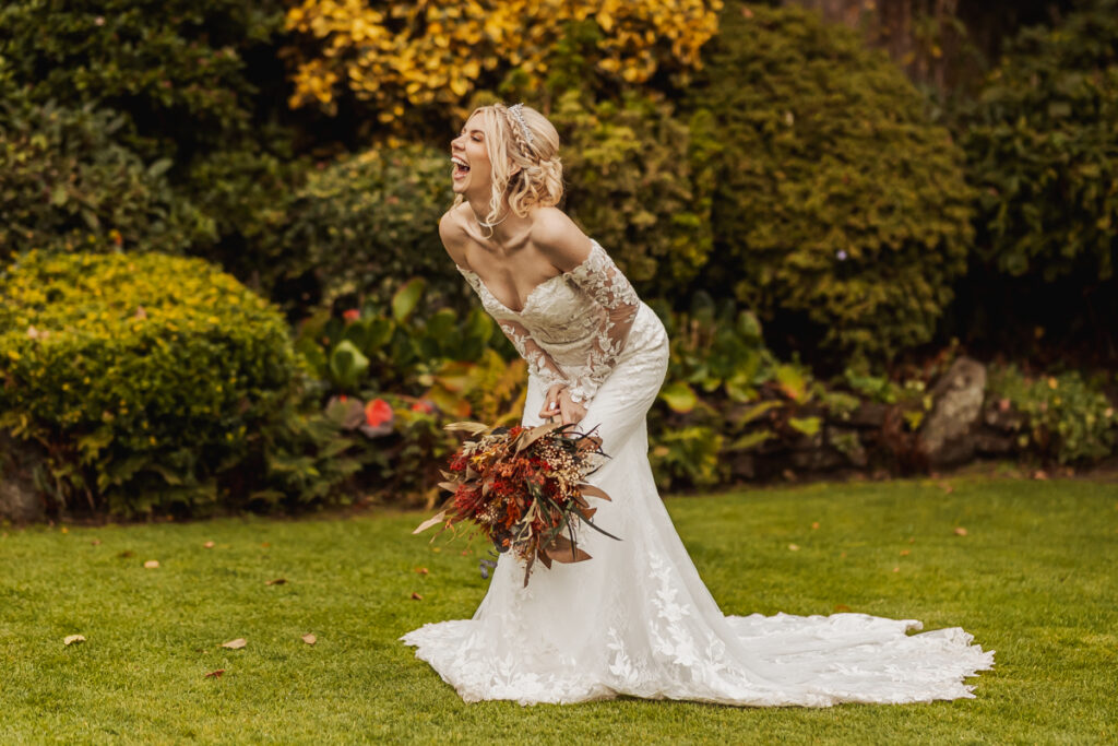 A bride in a white lace wedding gown laughs joyfully, holding a bouquet of autumn flowers at Ashes Barn, a charming Staffordshire wedding venue surrounded by lush greenery and golden foliage.