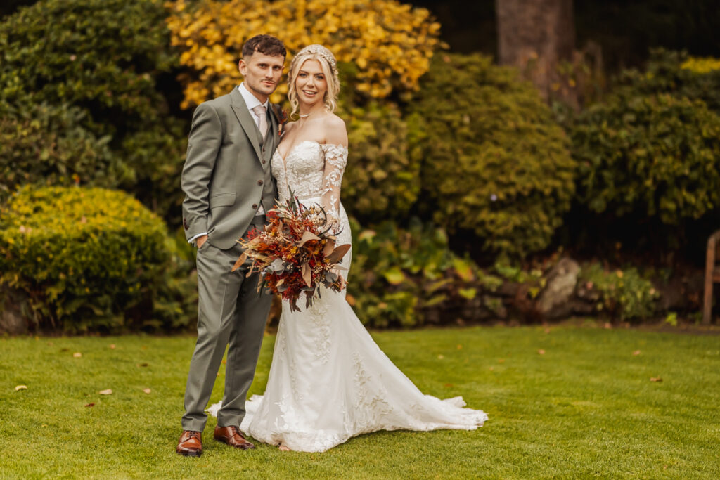 A bride in a white lace wedding dress and a groom in a gray suit stand together on green grass at Ashes Barn in Staffordshire, with the bride holding a bouquet of autumn flowers and lush greenery and yellow foliage in the background.