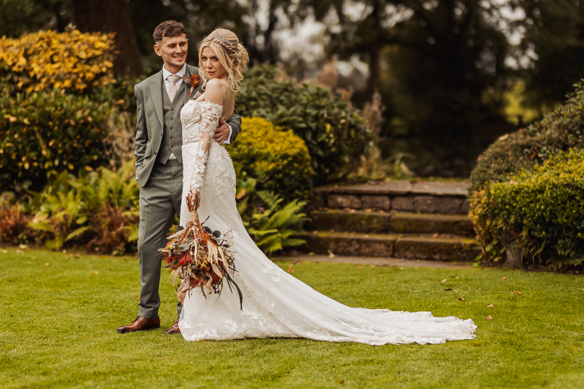 A bride in a long white dress with a floral bouquet stands next to a groom in a gray suit on a green lawn at Ashes Barn, surrounded by lush Staffordshire garden foliage and stone steps in the background.