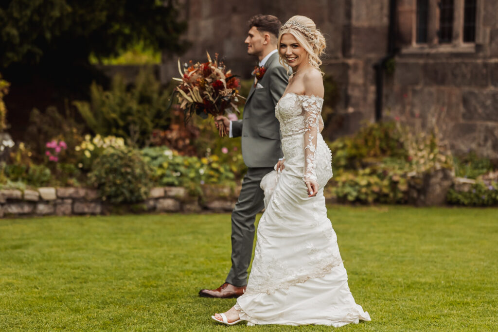 A bride in a white lace gown and a groom in a gray suit walk on the grass at Ashes Barn, Staffordshire; the bride smiles over her shoulder, holding her wedding dress, while the groom carries her bouquet.