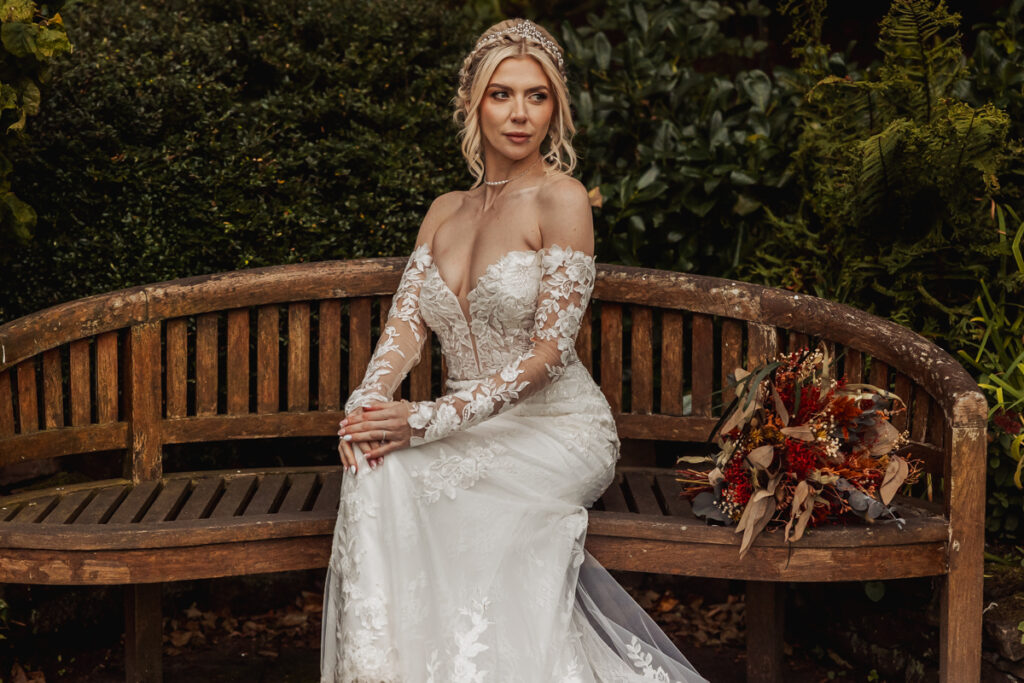 A bride in a long-sleeved, off-the-shoulder lace wedding dress sits on a wooden bench at Ashes Barn in Staffordshire, holding her hands in her lap, with an autumnal bouquet resting beside her.