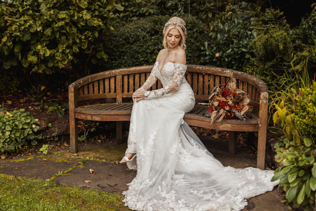 A bride in a long, white, lace wedding dress with sheer sleeves sits on a wooden bench at Ashes Barn in Staffordshire, holding her hands in her lap. A bouquet of red and orange flowers rests beside her, capturing the perfect wedding moment.