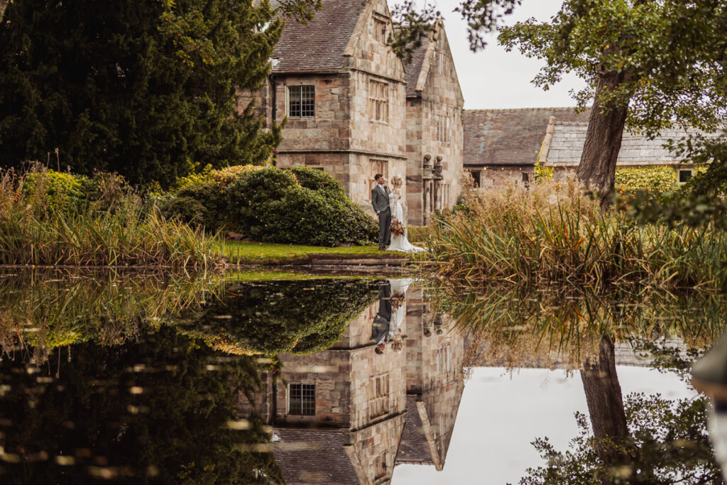 A bride and groom stand together on a grassy bank beside a pond at Ashes Barn in Staffordshire, with the historic stone building behind them. Their reflection, along with the building and trees, is clearly visible in the still water.