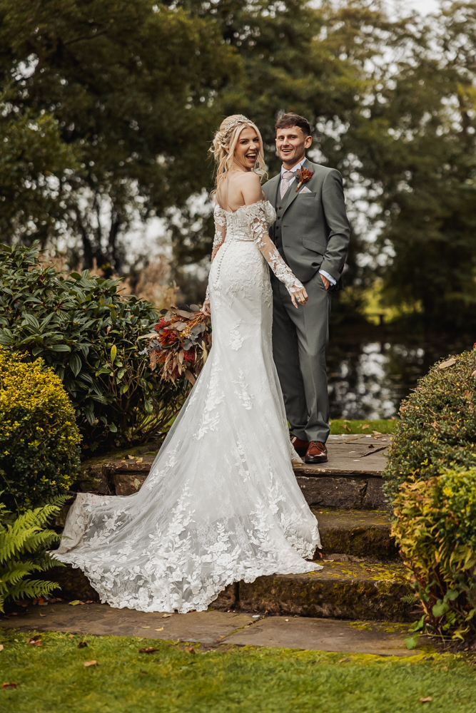 A bride in a white lace gown and a groom in a gray suit stand on stone steps outdoors at Ashes Barn in Staffordshire, holding hands and smiling, surrounded by greenery and trees.
