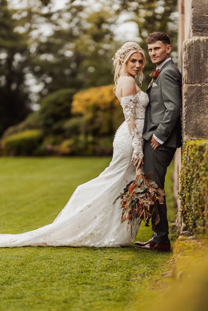 A bride in a lace wedding dress stands beside a groom in a dark suit near a stone wall at Ashes Barn Wedding in Staffordshire, holding a bouquet of dried flowers, with greenery and trees in the background.