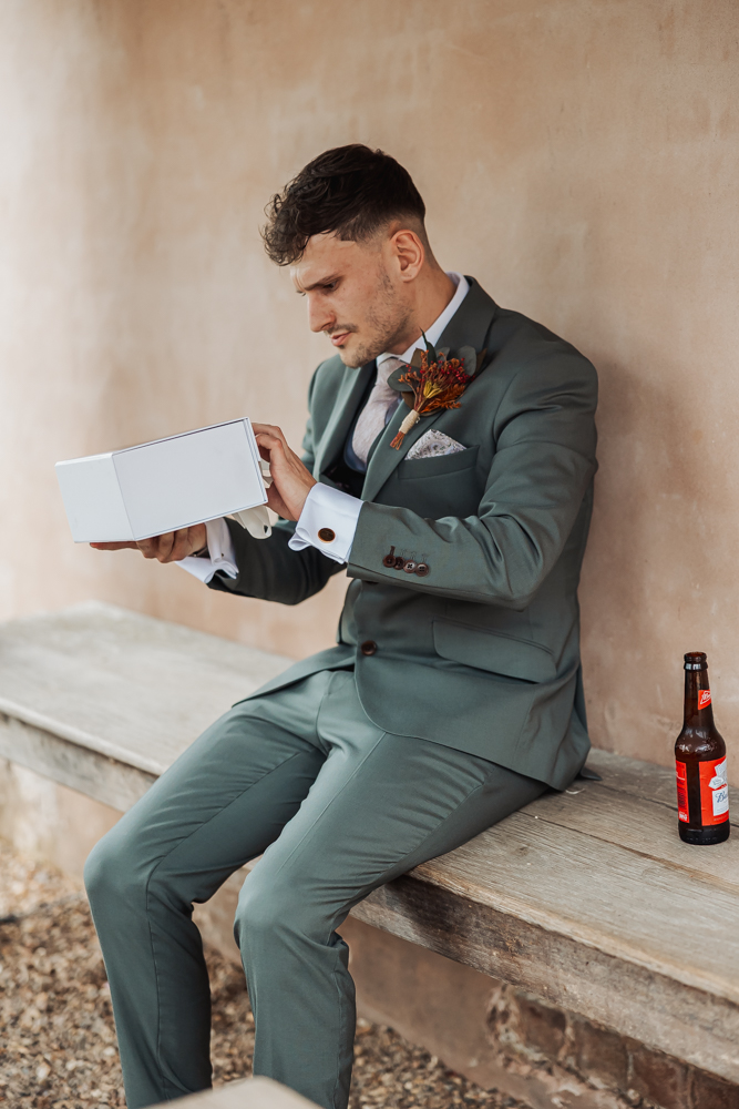 A man in a gray-green suit sits on a wooden bench at Ashes Barn, Staffordshire, opening a white box. He wears a boutonnière and dress shoes, with a beer bottle beside him. The background is a textured, neutral-colored wall.