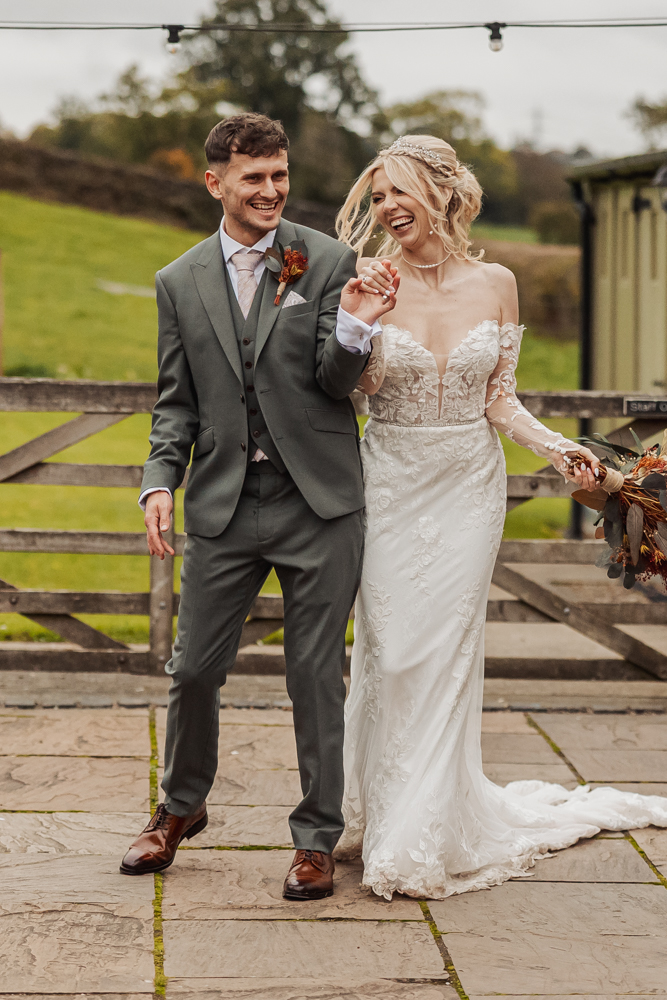 A smiling bride and groom walk hand-in-hand outdoors at Ashes Barn in Staffordshire. The bride wears a white lace off-shoulder dress and holds a bouquet, while the groom wears a green suit and brown shoes by a wooden fence and green field.