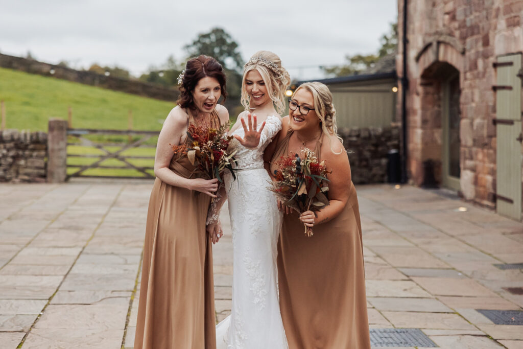 Three women stand outdoors on a stone patio at Ashes Barn in Staffordshire. The bride in a white dress shows off her ring while two bridesmaids in tan dresses, holding bouquets, lean in and smile. A rustic building and green field are behind them.