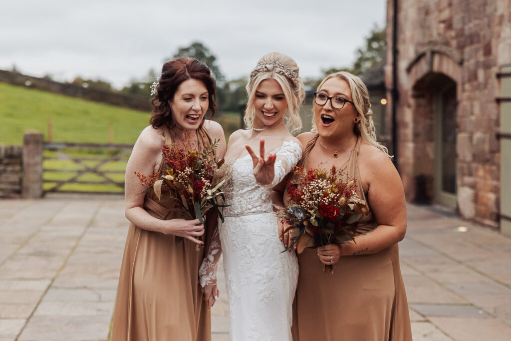 Three women in formal dresses stand outside Ashes Barn in Staffordshire. The bride in a white gown flashes a peace sign, smiling, while two bridesmaids in tan dresses laugh and hold bouquets of autumn flowers at this beautiful wedding.