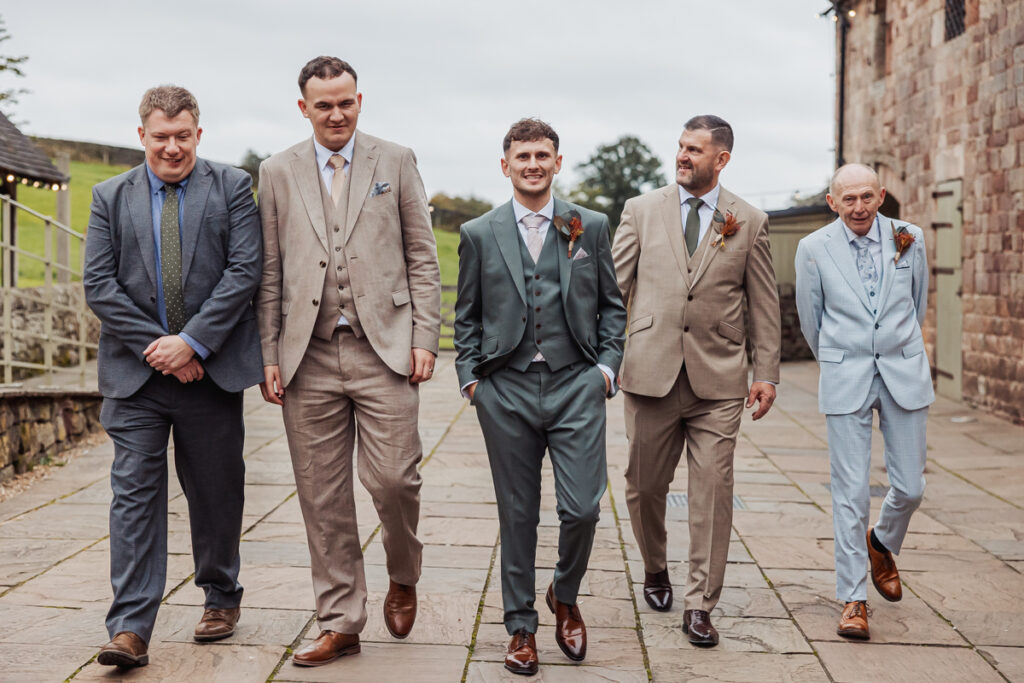 Five men in suits walk together on a stone path by a rustic building at Ashes Barn, Staffordshire, smiling and talking. Lush greenery and a cloudy sky suggest a wedding or celebratory occasion.