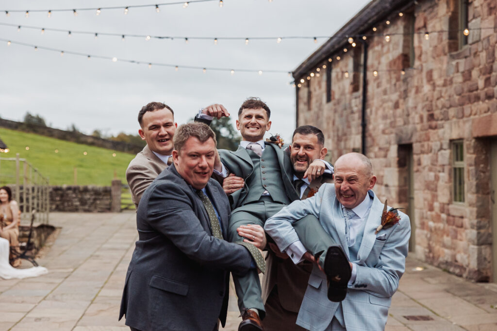 Five men in suits laugh together as they lift one of their friends off the ground outside Ashes Barn in Staffordshire, with string lights overhead, creating a joyful, celebratory wedding scene.