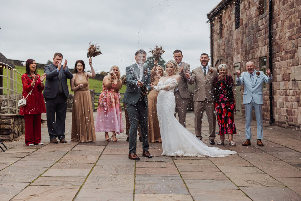 A bride and groom celebrate outdoors at Ashes Barn in Staffordshire by popping a bottle of champagne, surrounded by cheering friends and family in formal attire, with a stone building and cloudy sky in the background.