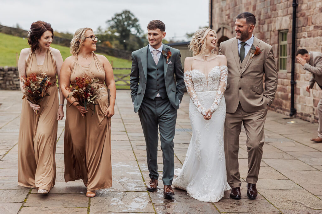 Four people, two in beige dresses holding bouquets and two men in suits, walk with a bride in a white gown outside Ashes Barn in Staffordshire. They are smiling and talking, with a stone building and greenery in the background.