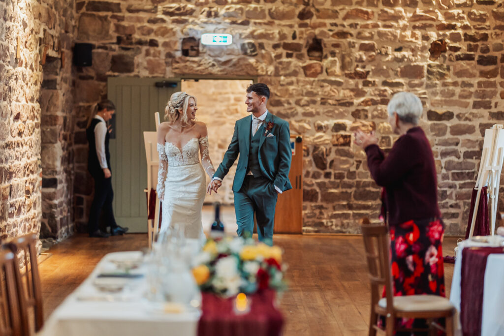 A bride and groom smile and hold hands while entering Ashes Barn, a rustic wedding venue in Staffordshire, as an older woman claps and a server stands by the door in the background.