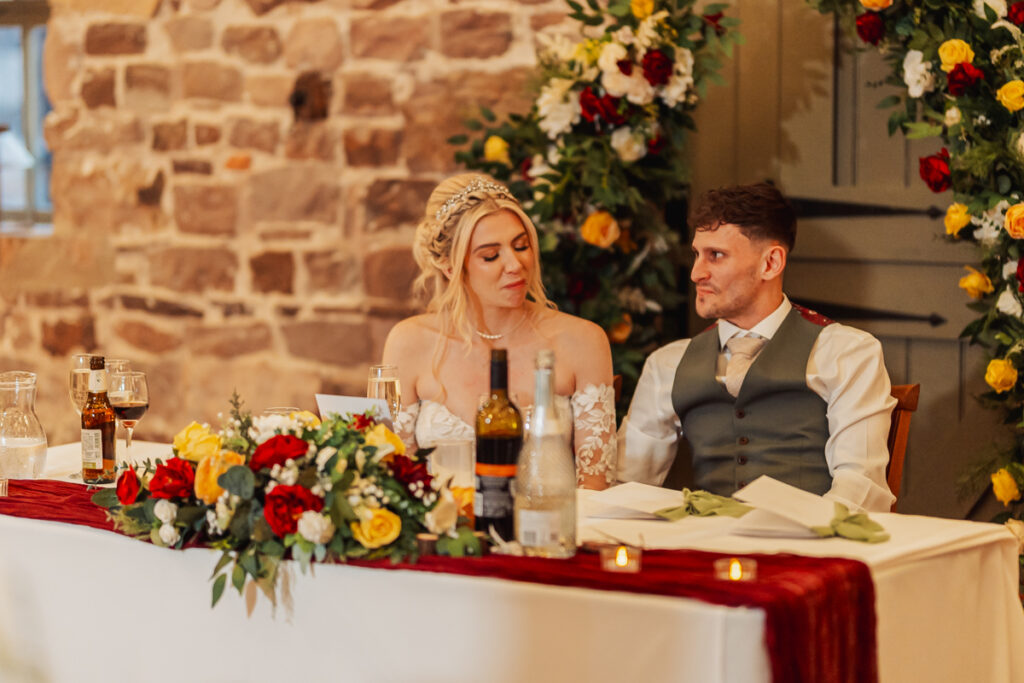 A bride and groom sit at a decorated table with red, yellow, and white flowers, bottles, and glasses in front of them. They look at each other, surrounded by floral arrangements and a rustic brick wall at Ashes Barn in Staffordshire.