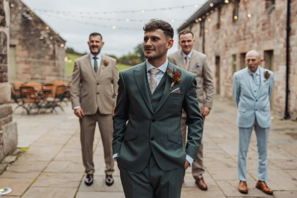 A groom in a green suit stands in front, hands in his pockets, with three groomsmen behind him in tan and light blue suits, outdoors between stone buildings with string lights above at a memorable Ashes Barn Wedding in Staffordshire.