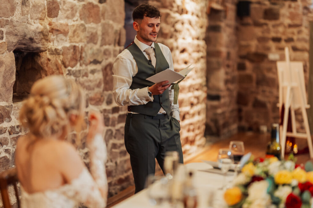 At Ashes Barn in Staffordshire, a man in a suit reads a heartfelt speech at a wedding reception, while a woman in a white dress listens from the beautifully decorated table in the rustic stone-walled venue.