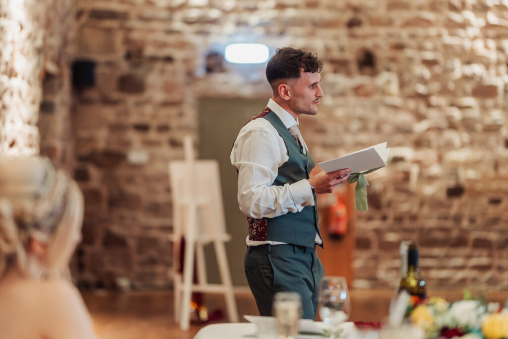 At Ashes Barn in Staffordshire, a man in a vest and tie stands holding a notebook, speaking at a wedding in a rustic brick-walled room. A blurred woman sits in the foreground, with wine and glasses on the table.