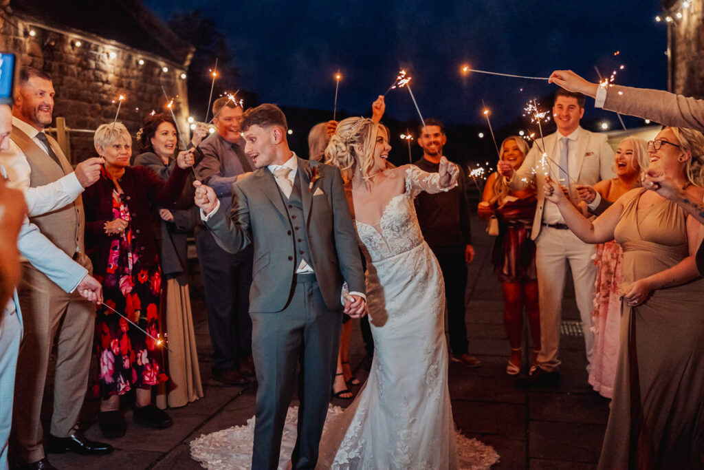 A newlywed couple holding hands and smiling walk through a crowd of guests with sparklers at night, celebrating their wedding outdoors at Ashes Barn in Staffordshire, with string lights and stone buildings in the background.