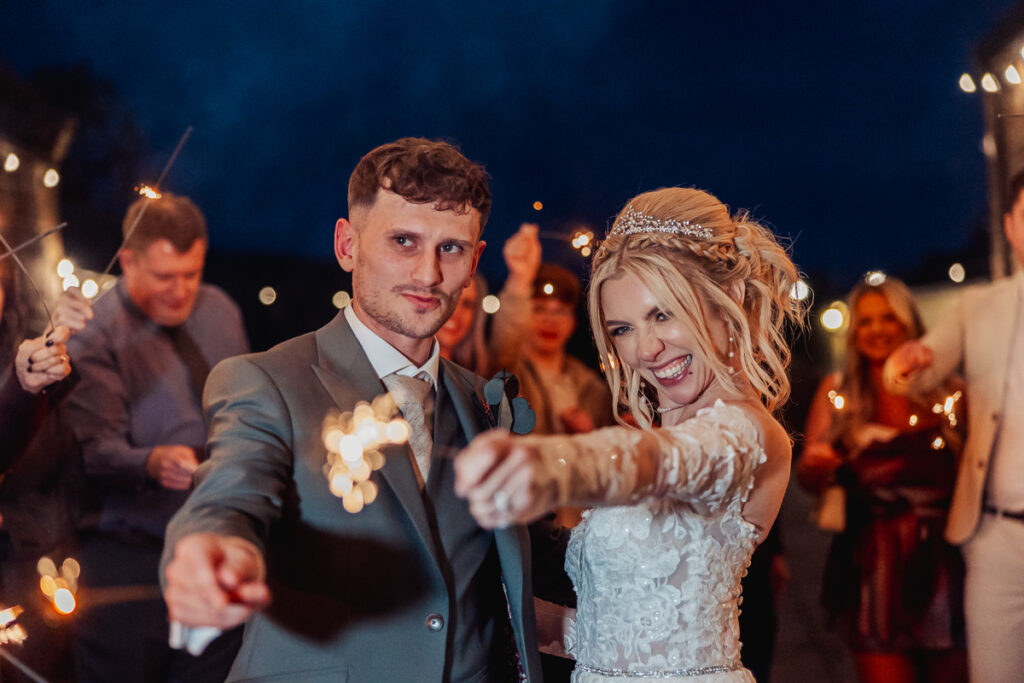 A bride and groom smile and hold sparklers toward the camera at night at Ashes Barn in Staffordshire, surrounded by wedding guests celebrating with sparklers in the background. The bride wears a white dress, and the groom is in a suit.