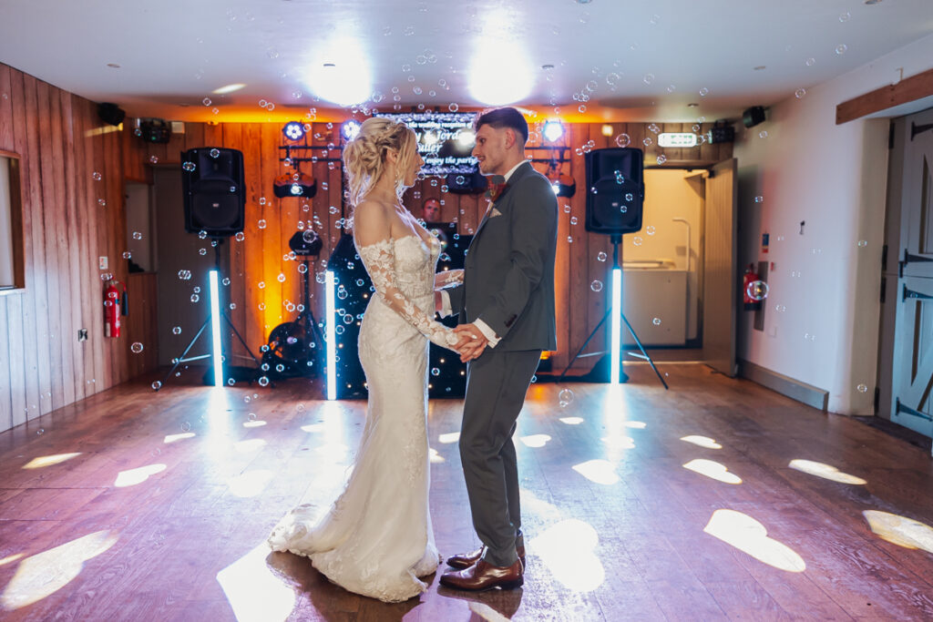 A bride and groom hold hands and gaze at each other while dancing in a softly lit room at Ashes Barn, Staffordshire, with bubbles floating around them. The bride wears a white gown and the groom is in a gray suit. Heart-shaped lights decorate the floor.