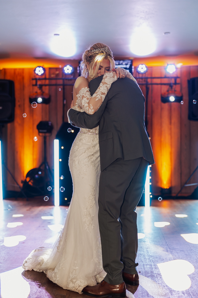 A bride in a white lace gown and a groom in a dark suit share an emotional embrace during their first dance at Ashes Barn Wedding venue in Staffordshire, surrounded by glowing lights and floating bubbles in a warmly lit setting.
