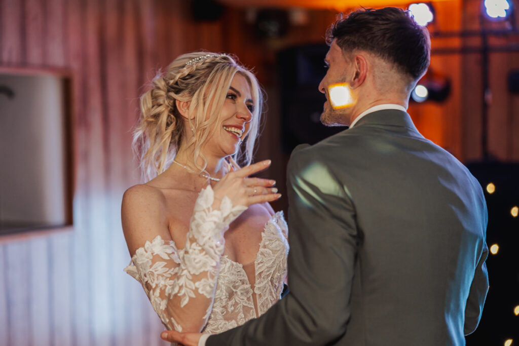 A bride in a white lace wedding gown and a groom in a suit smile and dance together indoors at Ashes Barn, Staffordshire, sharing a joyful moment during their wedding celebration. Warm lighting highlights their faces and expressions.