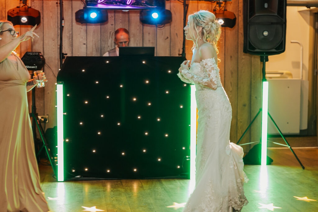A bride in a white lace dress dances and smiles under colorful lights near a DJ booth at Ashes Barn in Staffordshire, while a guest in a tan dress gestures with a drink in hand. Wooden paneling and glowing neon lights set the perfect wedding scene.