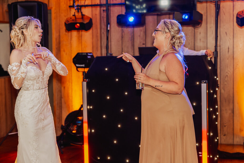 Two women, one in a white wedding dress and one in a tan dress, are dancing and chatting animatedly in front of a DJ booth with colorful lights at an indoor Ashes Barn Wedding in Staffordshire.
