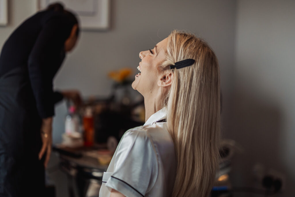 A blonde woman with hair clips smiles and laughs while sitting in a silky white top at Ashes Barn, Staffordshire. In the background, another person prepares items on a table, capturing a relaxed and candid wedding moment indoors.
